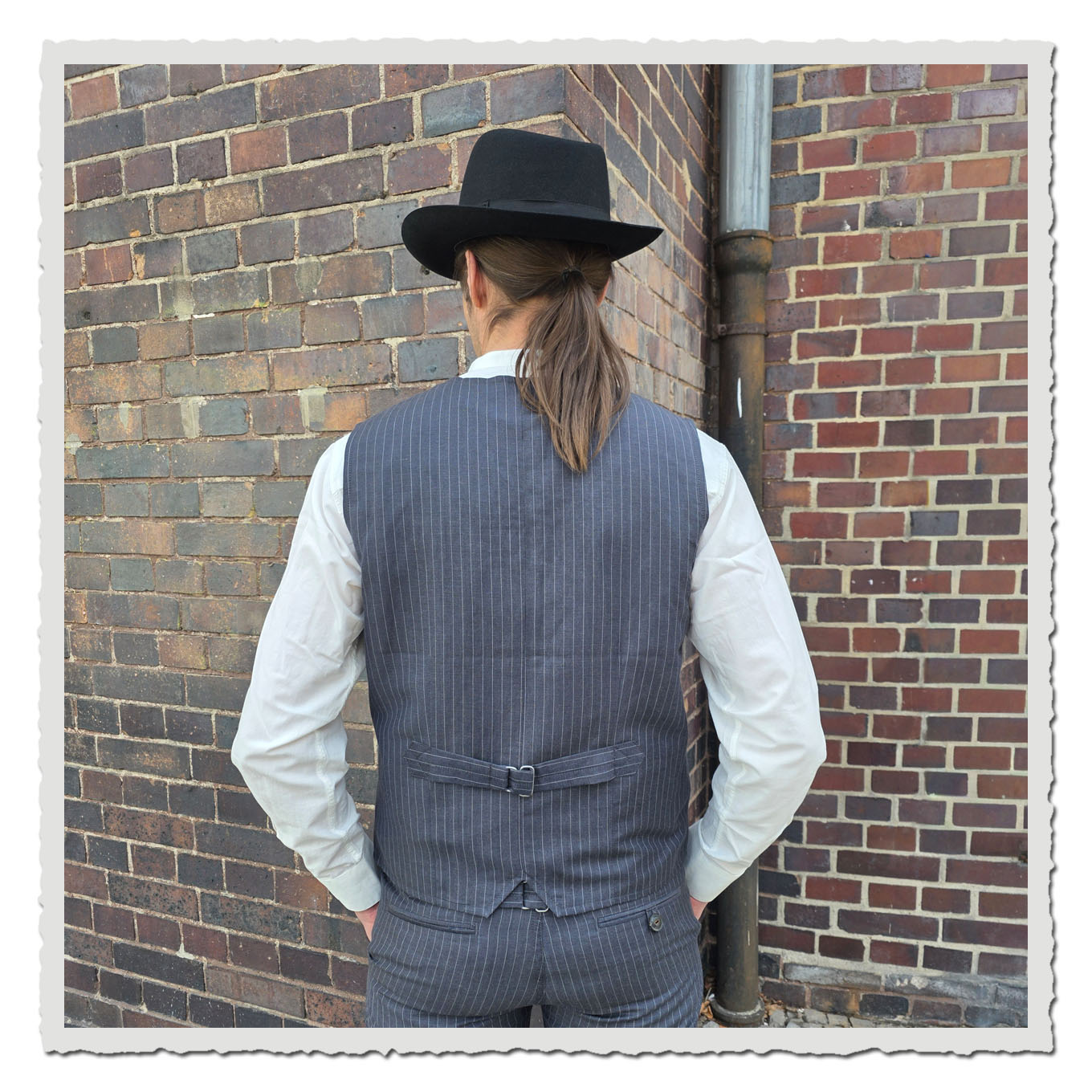 Rückansicht einer grauen Nadelstreifenweste am Model vor Backsteinwand im Außenbereich.